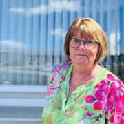 Die Frau mit kurzen Haaren und einer Brille lächelt leicht, während sie vor einem hellen Hintergrund mit Fenstern steht.