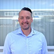 Der Mann trägt ein hellblaues Hemd und lächelt, während er vor einer Glasfassade mit reflektierenden Fenstern posiert.