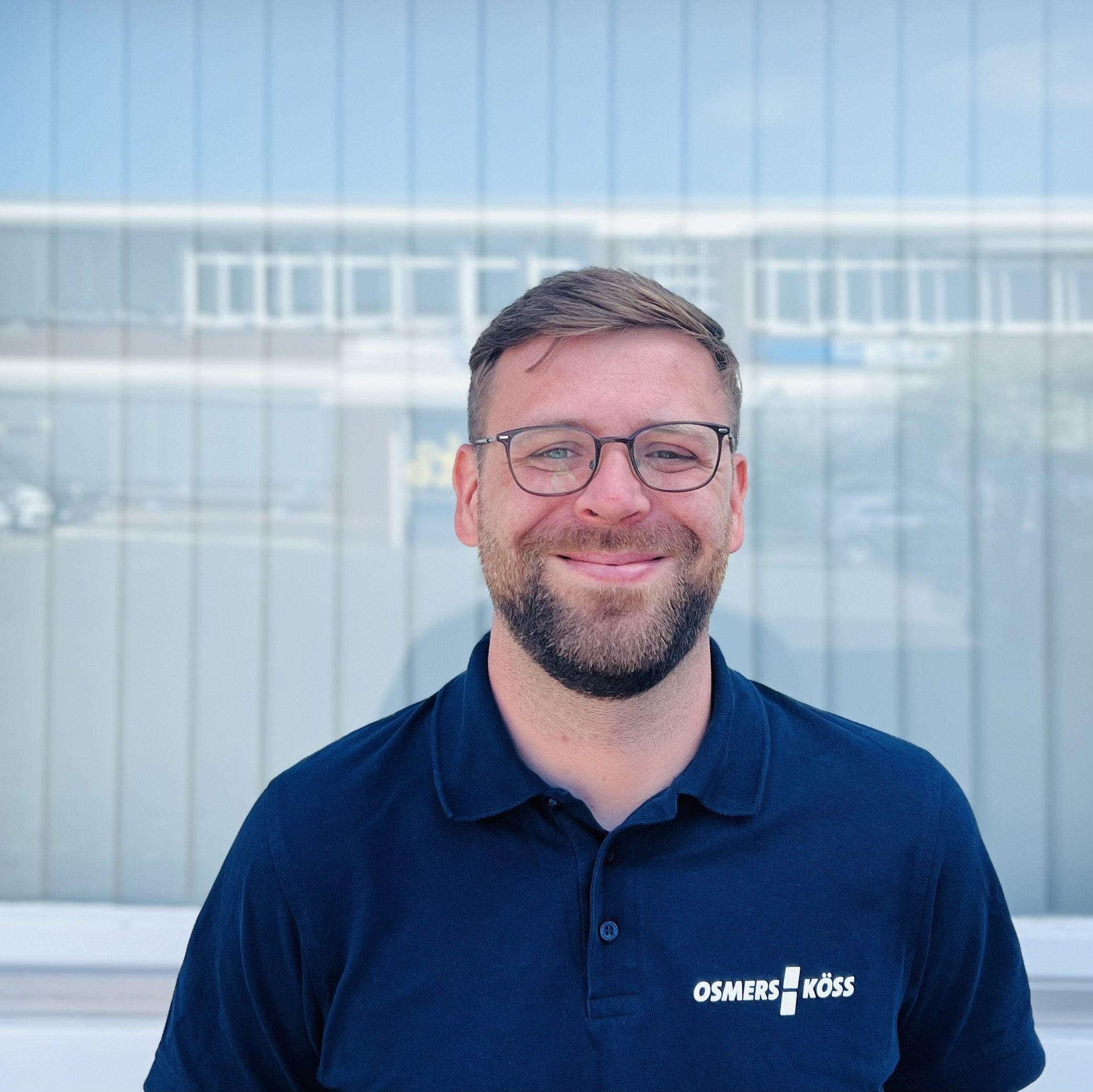 Der Mann trägt ein dunkelblaues Poloshirt mit dem Schriftzug OSMERS KÖSS und lächelt freundlich in die Kamera.