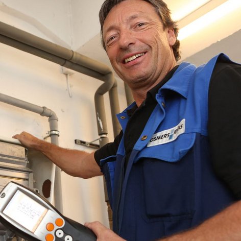 Der Handwerker zeigt auf eine Heizungsanlage, während er ein digitales Messgerät zur Überprüfung der Werte benutzt.