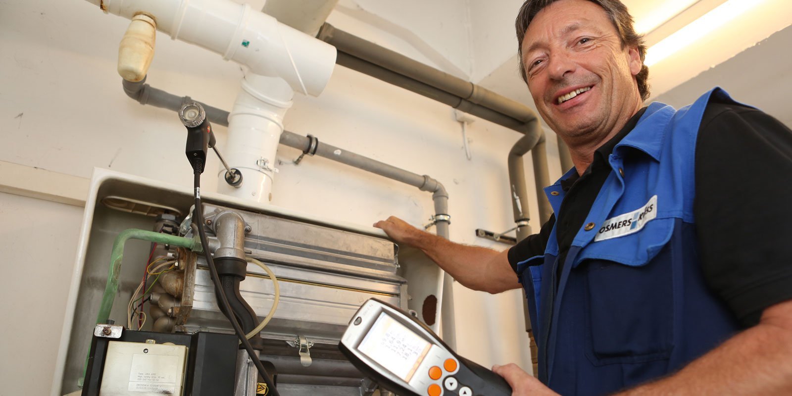 Der Handwerker zeigt auf eine Heizungsanlage, während er ein digitales Messgerät zur Überprüfung der Werte benutzt.