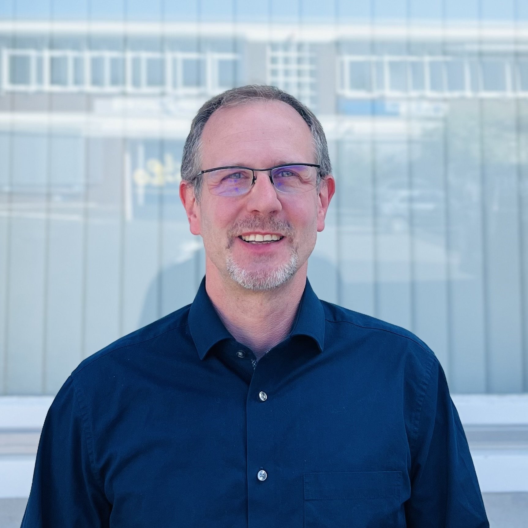 Der Mann trägt ein schwarzes Hemd und lächelt freundlich, während er vor einer Glasfassade posiert.