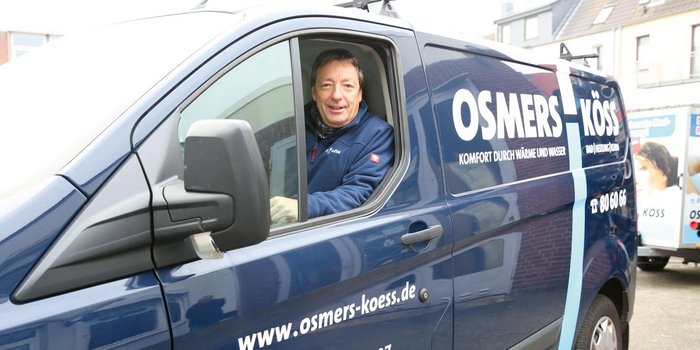 Der Fahrer eines OSMERS-KÖSS-Fahrzeugs lächelt, während er in einer urbanen Umgebung auf dem Fahrersitz sitzt.