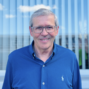 Ein Mann mit Brille und grauen Haaren lächelt vor einem modernen Gebäude mit dem Schriftzug 'OSMERS KÖSS'.