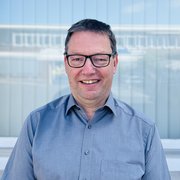 Vor einem großen Fenster mit dem Logo 'OSMERS KÖSS' steht ein Mann, der fröhlich in die Kamera lächelt.